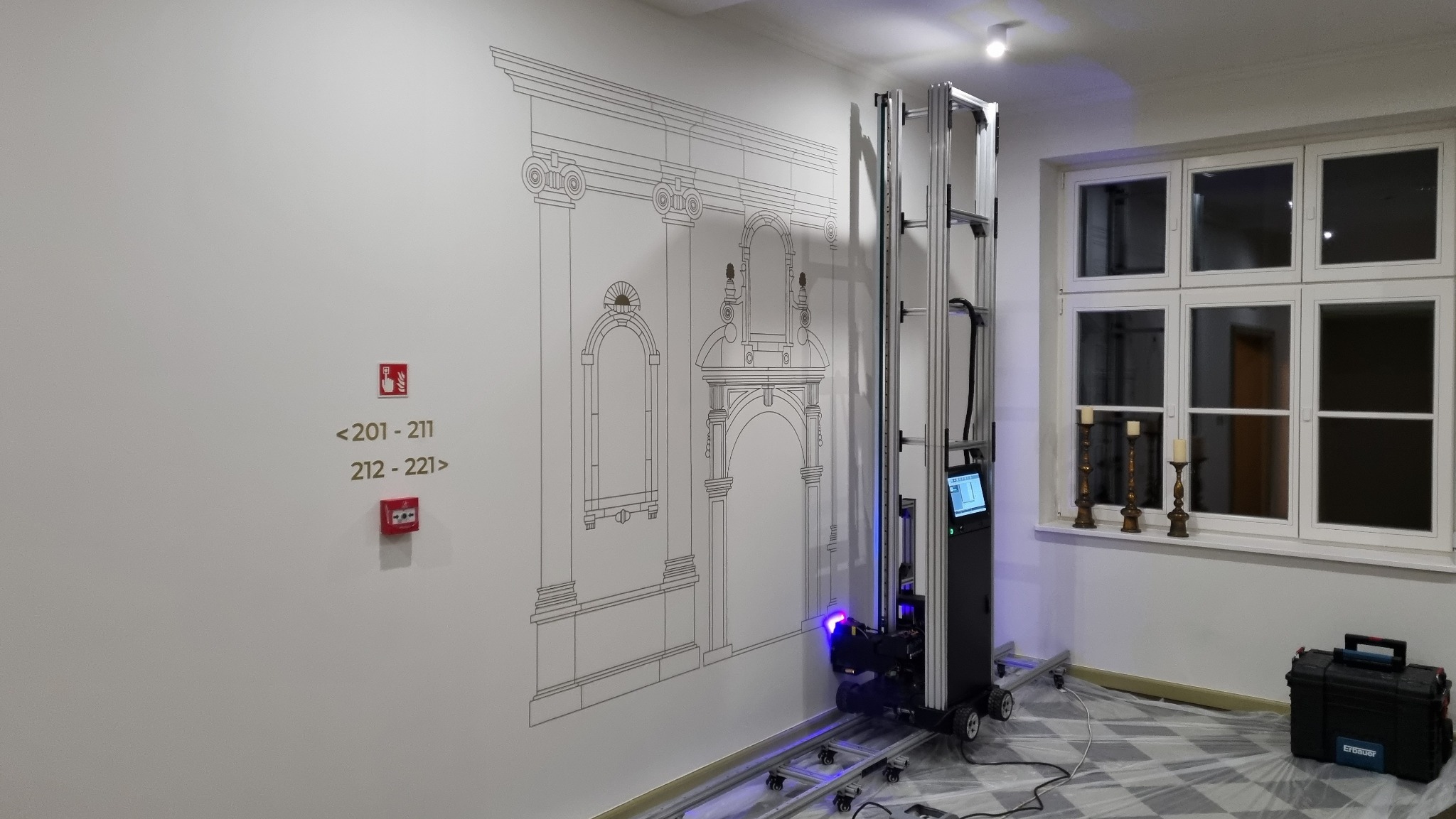 Wnętrze z maszyną do druku pionowego, nanoszącą na ścianę graficzny wzór architektoniczny w kolorze złotym. Obok okno z świecami, na podłodze folia ochronna.