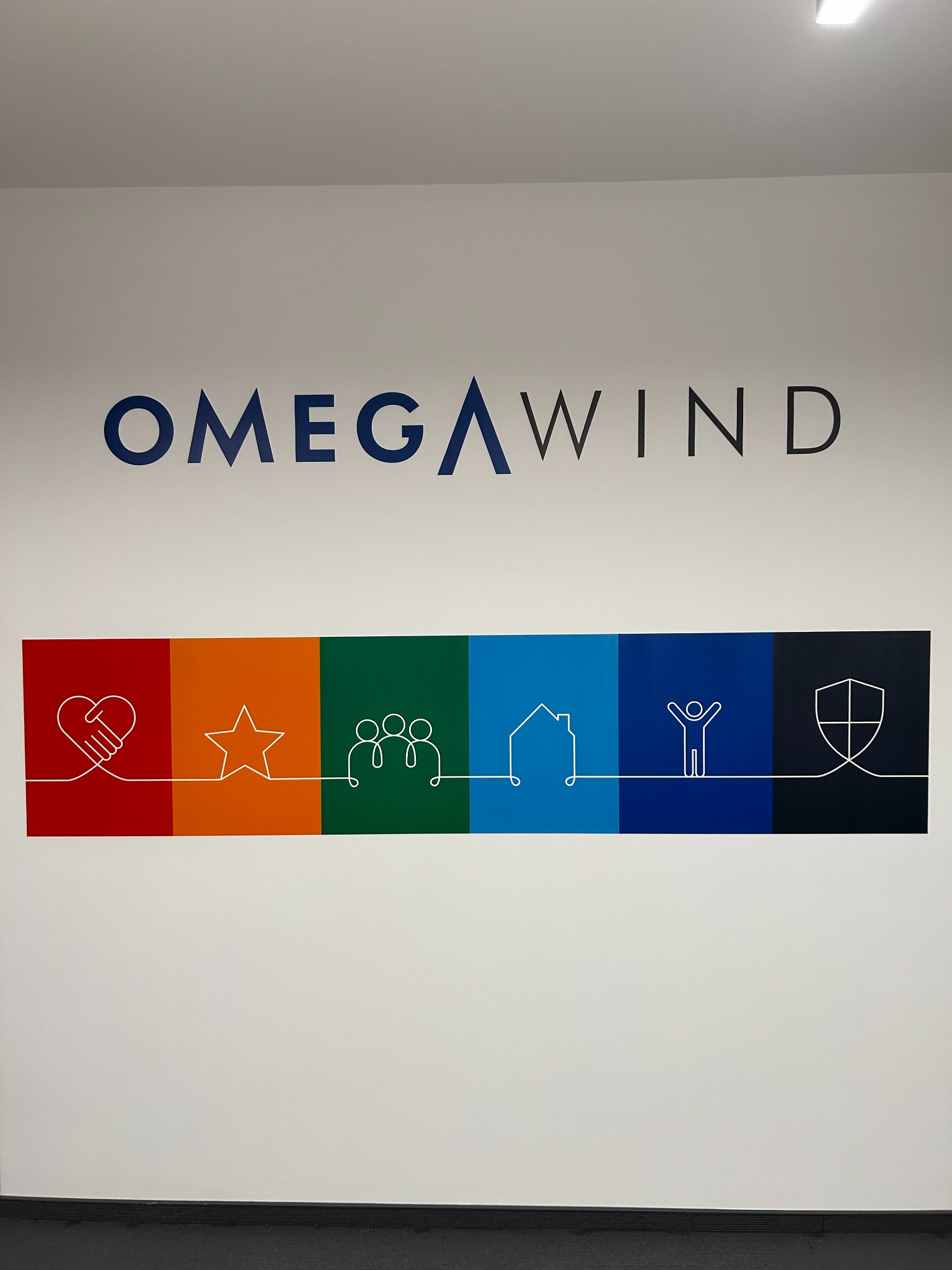 Ściana biura z logo Omega Wind i kolorowymi symbolami: serce, gwiazda, ludzie, dom, postać, tarcza. Minimalistyczny design, nowoczesne wnętrze.