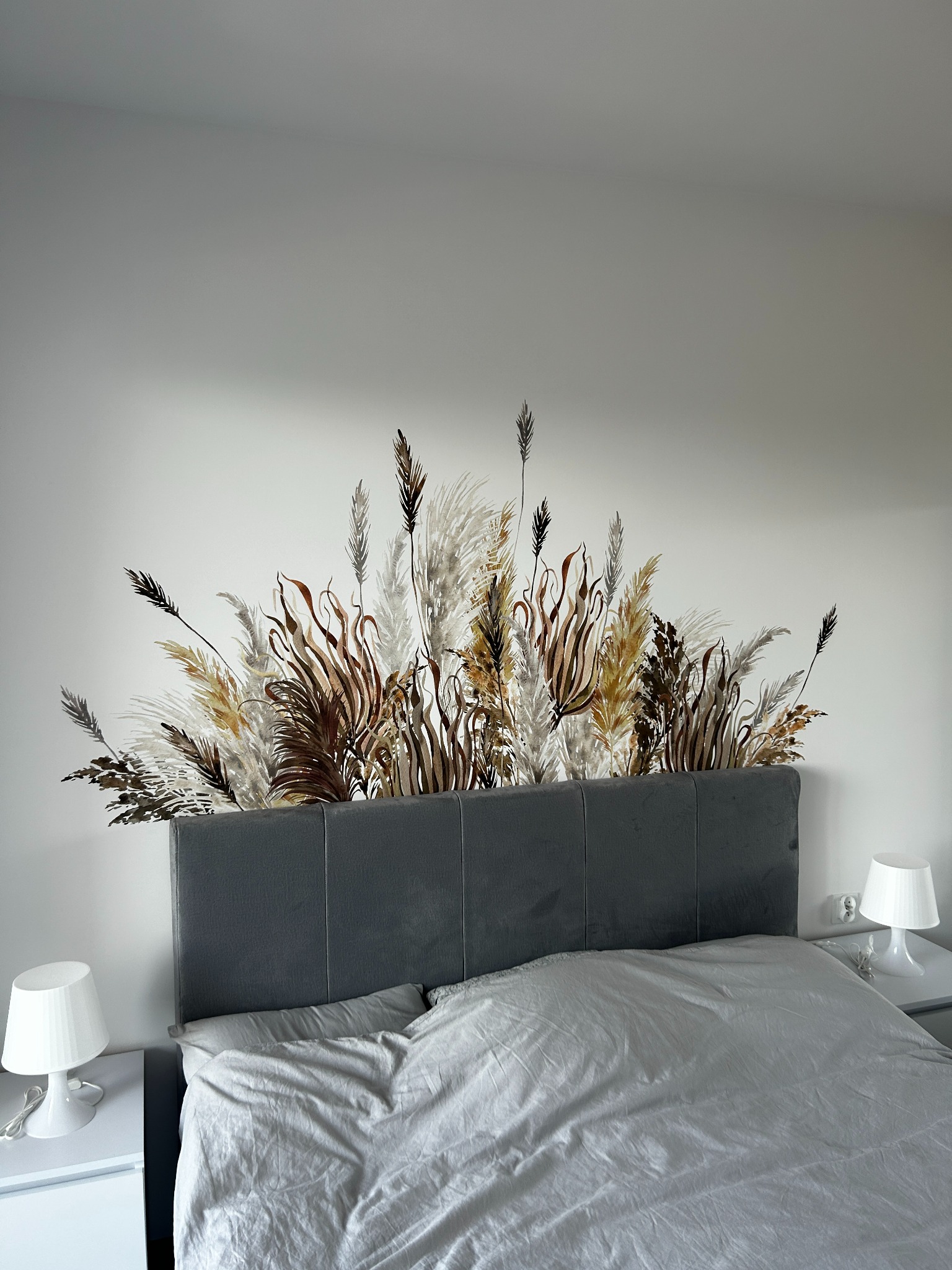 Sypialnia z szarym łóżkiem i naklejką ścienną z motywem roślinnym nad wezgłowiem. Minimalistyczne lampki nocne na białych szafkach.
