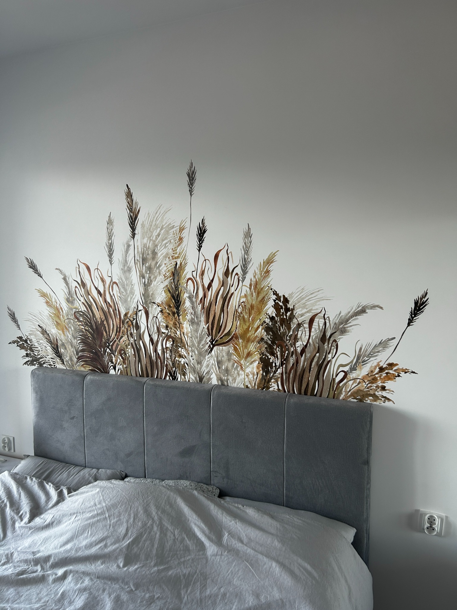Ściana w sypialni z dekoracyjnym malunkiem przedstawiającym trawy pampasowe nad wezgłowiem łóżka. Ujęcie z bliska, stonowana kolorystyka.