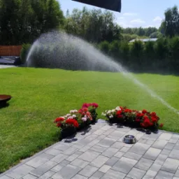 Automatyczne nawadnianie trawnika zraszaczem sektorowym. Widok na soczystą, zieloną trawę, kwiaty w donicach i palenisko ogrodowe w słoneczny dzień.