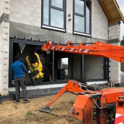 Vibest Home Sp. z o.o. - Montaż okna z użyciem dźwigu przyssawkowego na budowie domu z białego betonu komórkowego. Pracownik w ubraniu roboczym kieruje procesem.