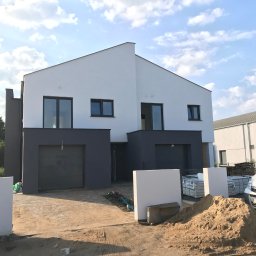 Vibest Home Sp. z o.o. - Nowoczesny bliźniak z szarymi bramami garażowymi i oknami, jasna elewacja, błękitne niebo z chmurami w tle. Widoczny piasek i auto dostawcze.