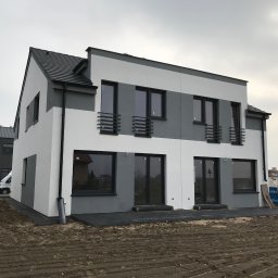 Vibest Home Sp. z o.o. - Dwukondygnacyjny bliźniak z szaro-białą elewacją, czarnymi oknami i balkonami z barierkami, widok z zewnątrz na plac budowy.