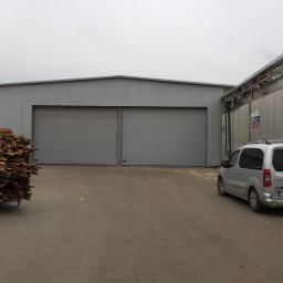 Vibest Home Sp. z o.o. - Szara hala magazynowa z dwuskrzydłowymi bramami. Na pierwszym planie palety z drewnem i zaparkowany srebrny samochód dostawczy.