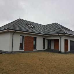 Vibest Home Sp. z o.o. - Nowoczesny dom jednorodzinny z grafitową bramą garażową, brązowymi akcentami elewacji i ciemną dachówką pod pochmurnym niebem. Widok z poziomu trawnika.