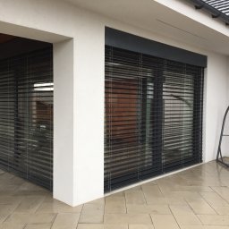 Vibest Home Sp. z o.o. - Eleganckie, grafitowe rolety zewnętrzne zamontowane na nowoczesnym budynku z białymi ścianami. Widoczne poziome lamele i fragment tarasu z jasnej kostki.