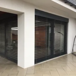 Eleganckie, grafitowe rolety zewnętrzne zamontowane na nowoczesnym budynku z białymi ścianami. Widoczne poziome lamele i fragment tarasu z jasnej kostki.