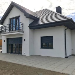 Vibest Home Sp. z o.o. - Elewacja domu jednorodzinnego z szarymi roletami zewnętrznymi na parterze i balkonem na piętrze, w tle błękitne niebo z chmurami.