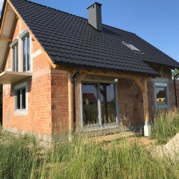 Vibest Home Sp. z o.o. - Surowy stan domu z cegły z zamontowanymi oknami i czarnym dachem, otoczony wysoką trawą i stertą piasku, ukazujący etap budowy z elementami wykończeniowymi.