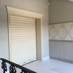 Vibest Home Sp. z o.o. - Beżowa brama rolowana w eleganckiej wnęce z dekoracyjną listwą, widoczna z perspektywy klatki schodowej, w jasnym wnętrzu z ozdobnymi płytkami.