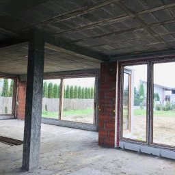 Vibest Home Sp. z o.o. - Surowy stan budynku z zamontowanymi oknami PCV, widoczne cegły i betonowy strop. Duże okna z widokiem na zieleń, wnętrze w trakcie budowy.