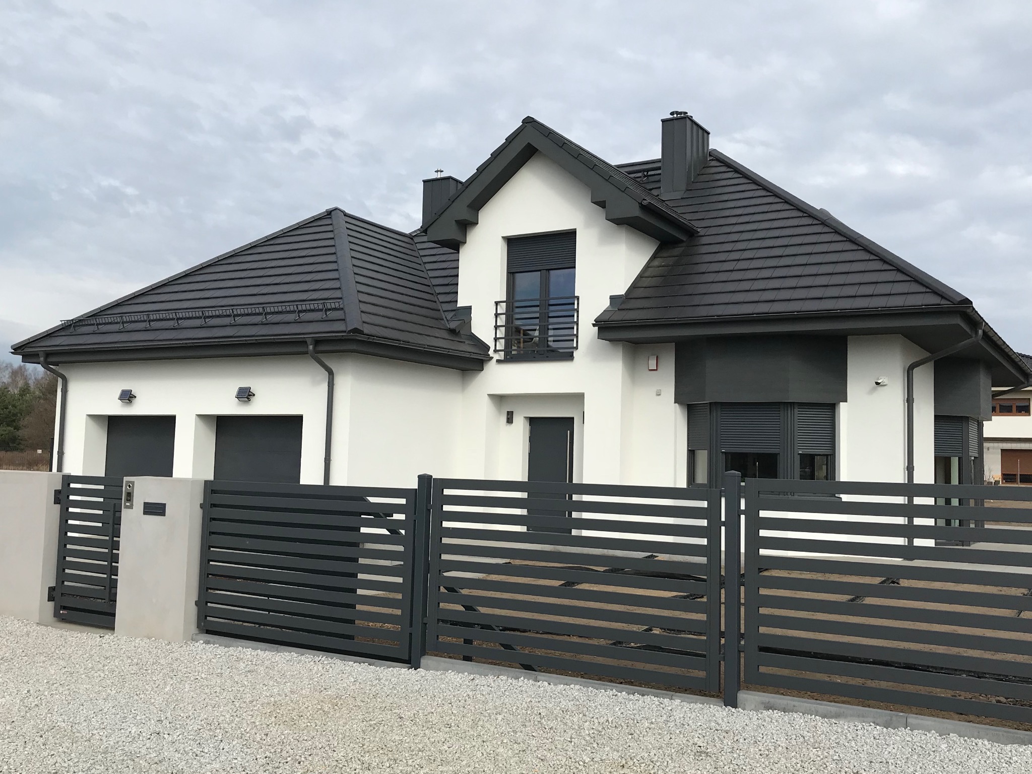 Nowoczesny dom z grafitową bramą poziomą, roletami zewnętrznymi i dwuspadowym dachem z ciemnej dachówki. Minimalistyczny styl, jasna elewacja, żwirowe podłoże.