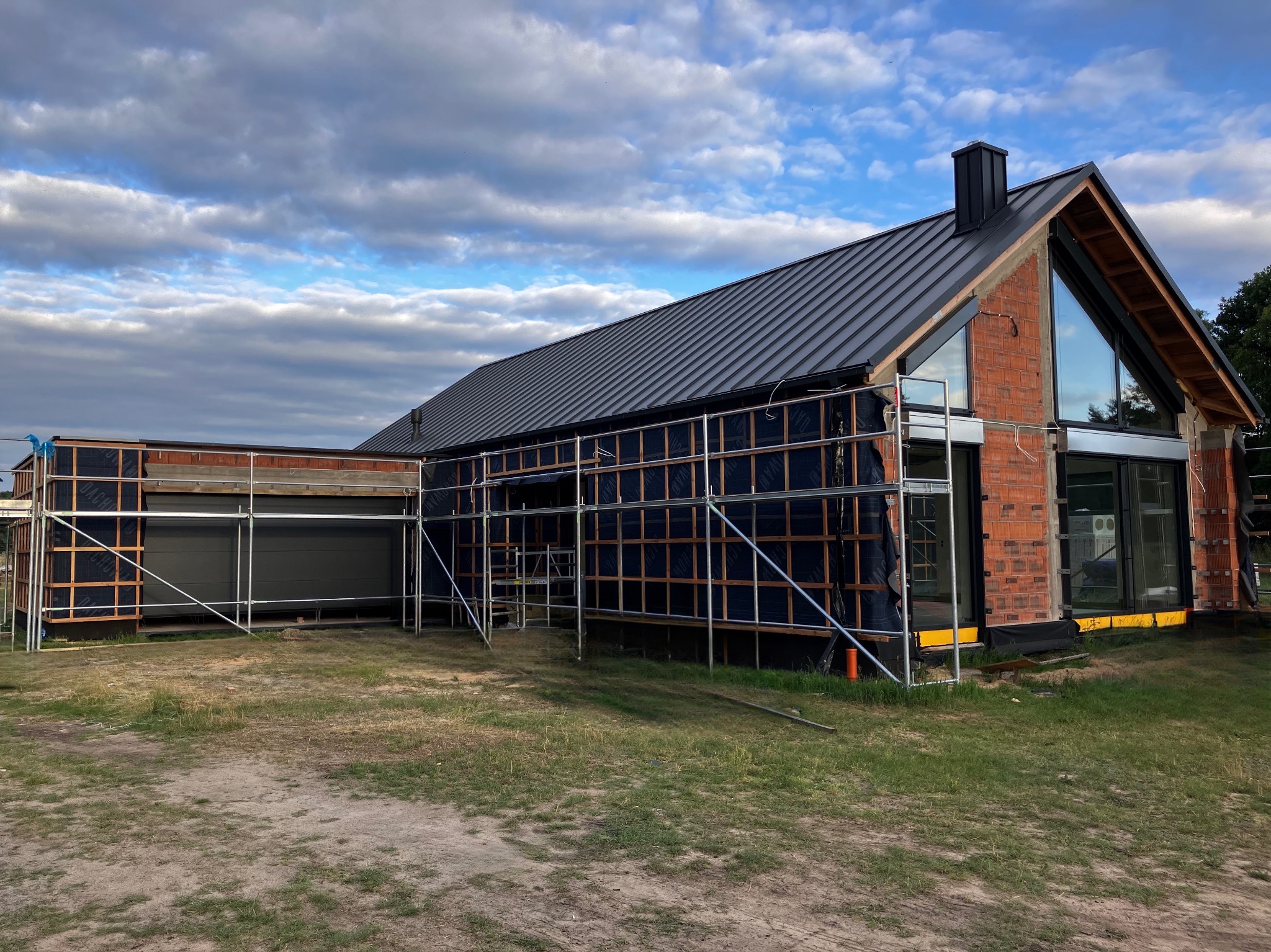 Budowa domu jednorodzinnego z cegły, z widocznymi rusztowaniami i zamontowanymi oknami. W tle pochmurne niebo.