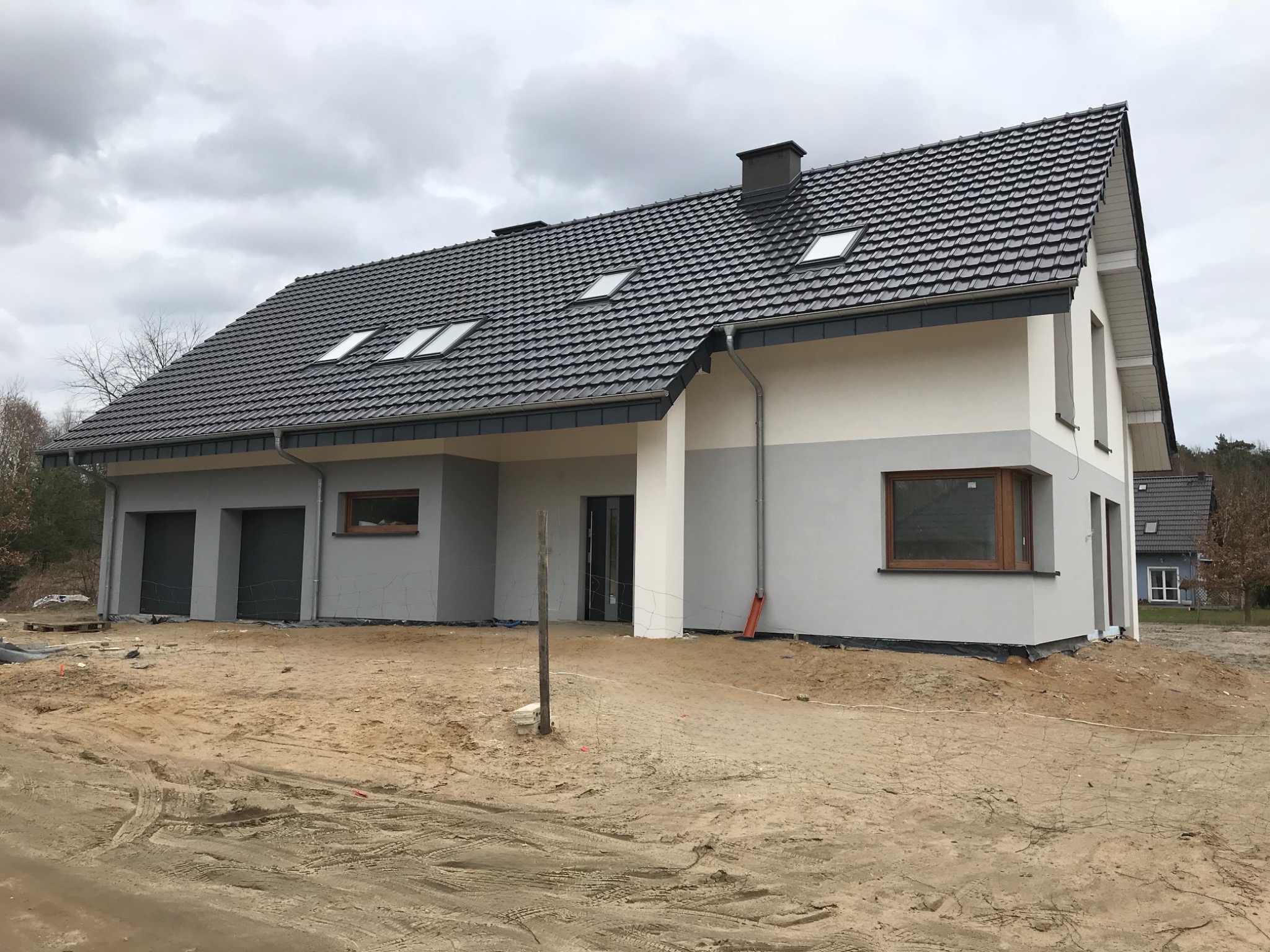 Nowoczesny dom jednorodzinny z szarą dachówką, oknami połaciowymi i dwukolorową elewacją w trakcie wykończenia, na działce z piaskiem.