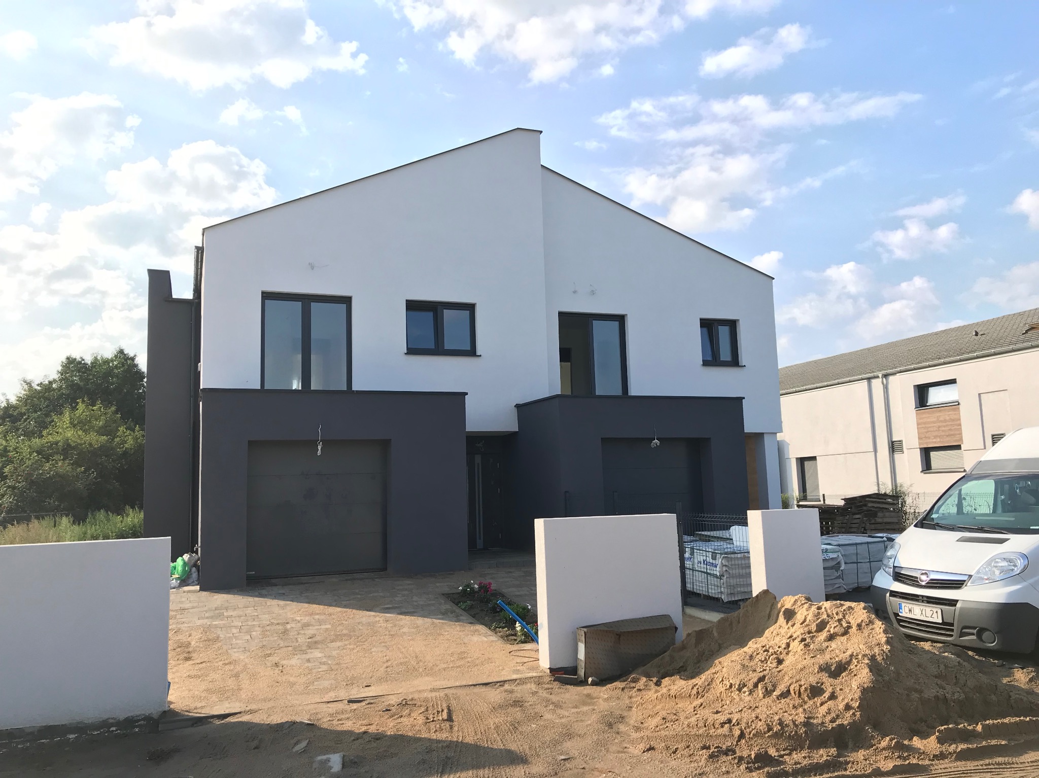 Nowoczesny bliźniak z szarymi bramami garażowymi i oknami, jasna elewacja, błękitne niebo z chmurami w tle. Widoczny piasek i auto dostawcze.