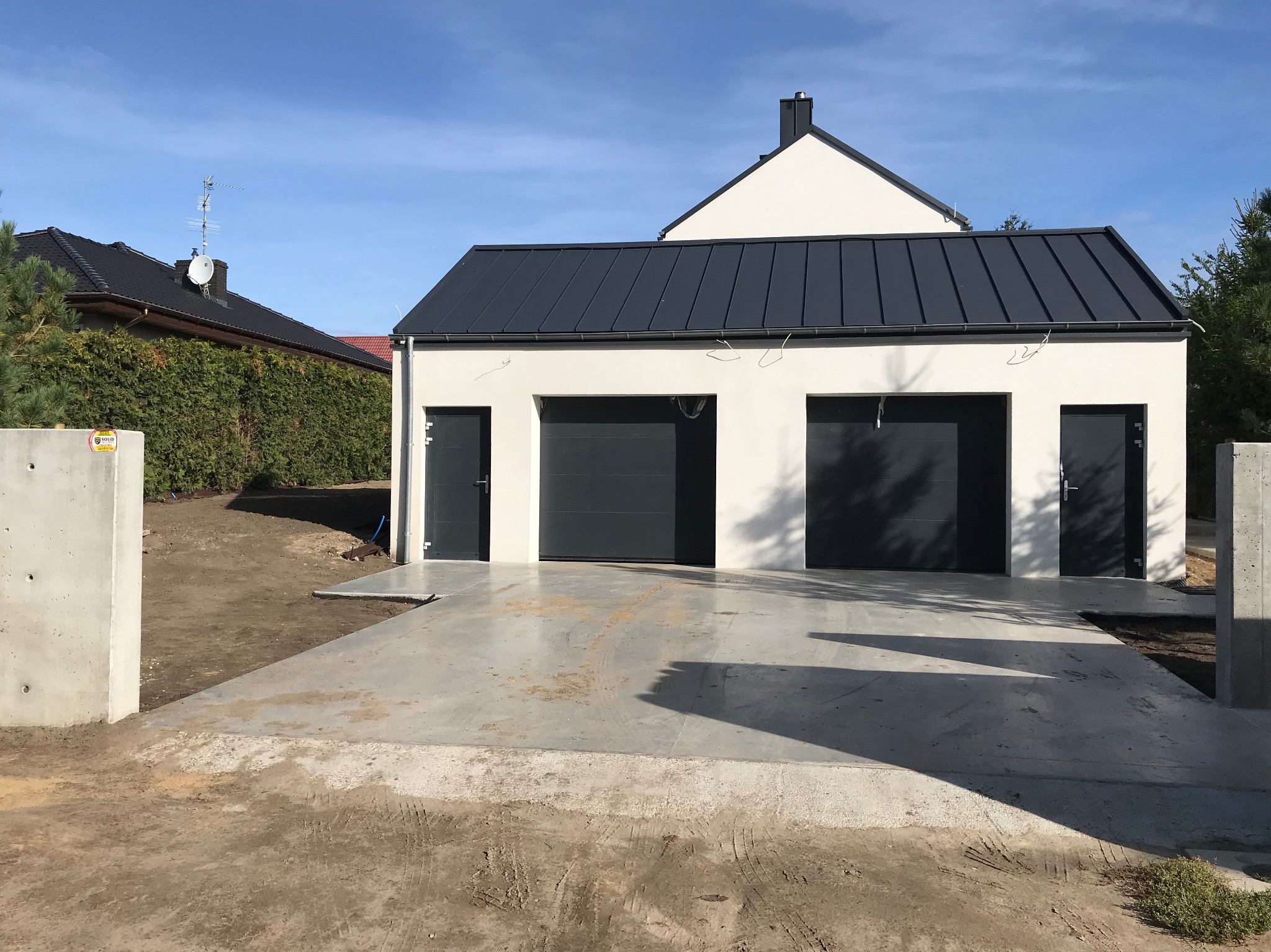 Nowoczesny garaż z dwoma bramami w kolorze antracytu i dodatkowymi drzwiami wejściowymi. Jasna elewacja, ciemny dach. Betonowy podjazd.