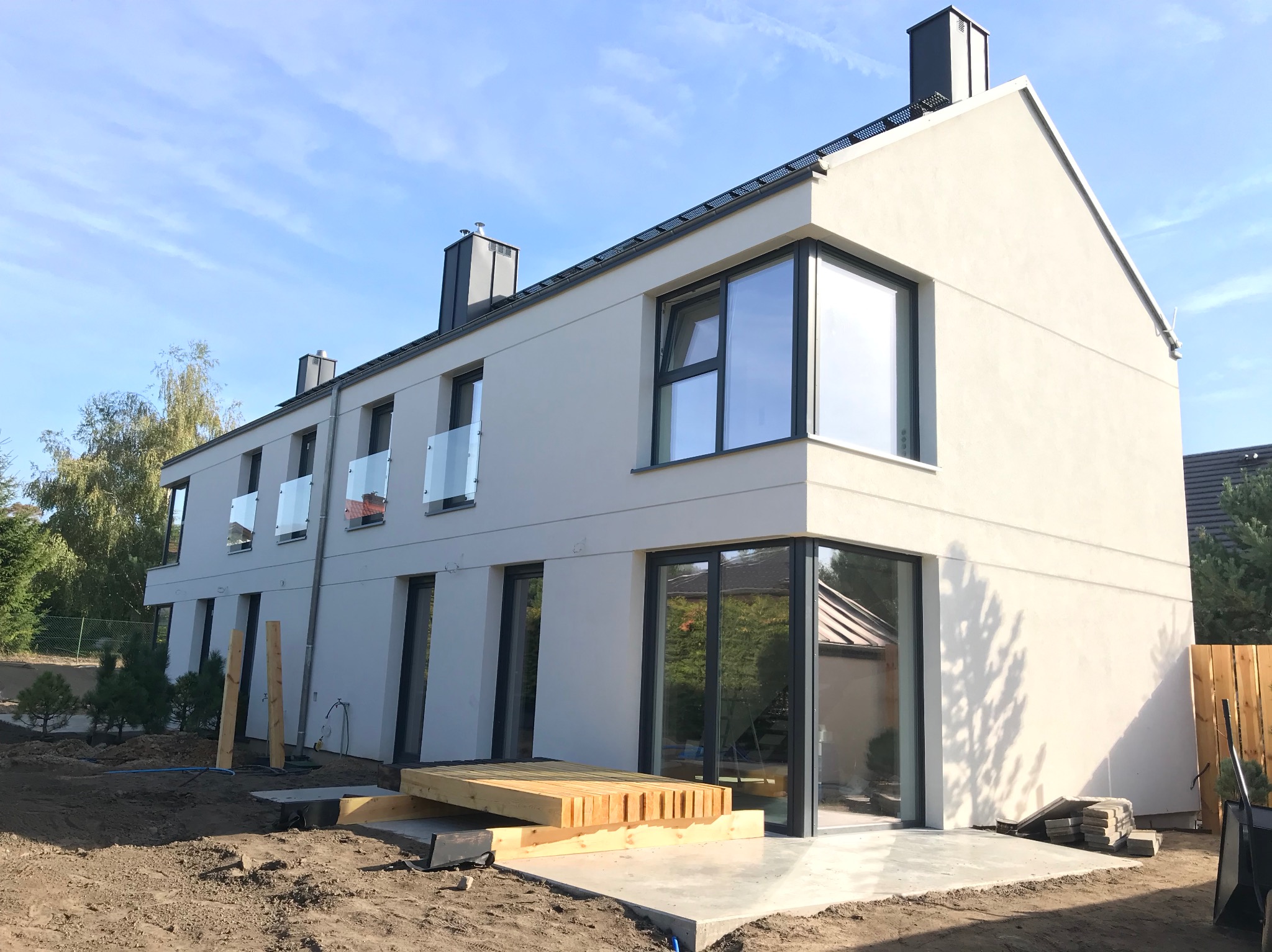 Nowoczesny, trzypiętrowy dom z dużymi oknami i szklanymi barierkami balkonowymi. Widoczny fragment tarasu w trakcie budowy i świeżo posadzona zieleń.