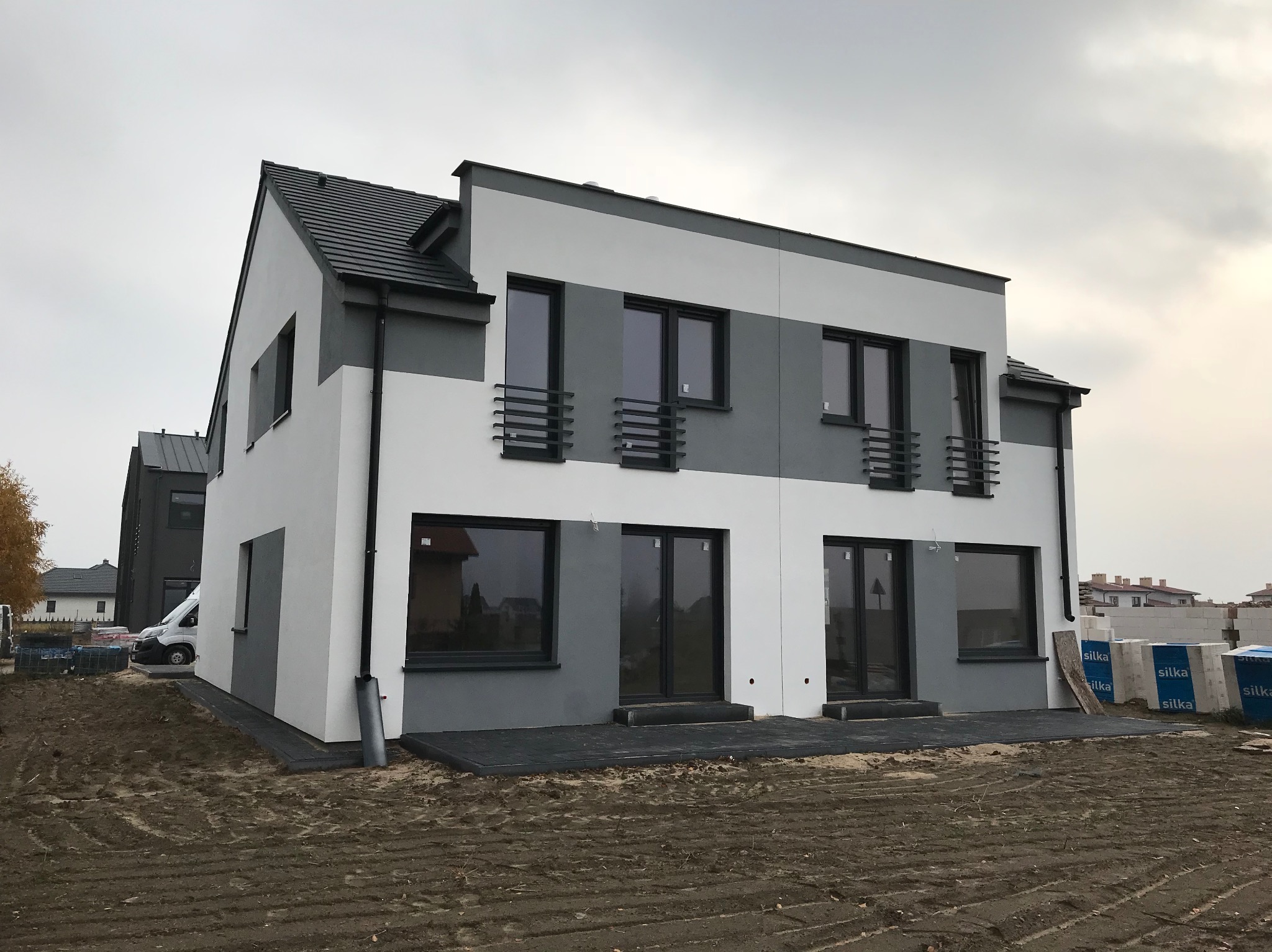 Dwukondygnacyjny bliźniak z szaro-białą elewacją, czarnymi oknami i balkonami z barierkami, widok z zewnątrz na plac budowy.