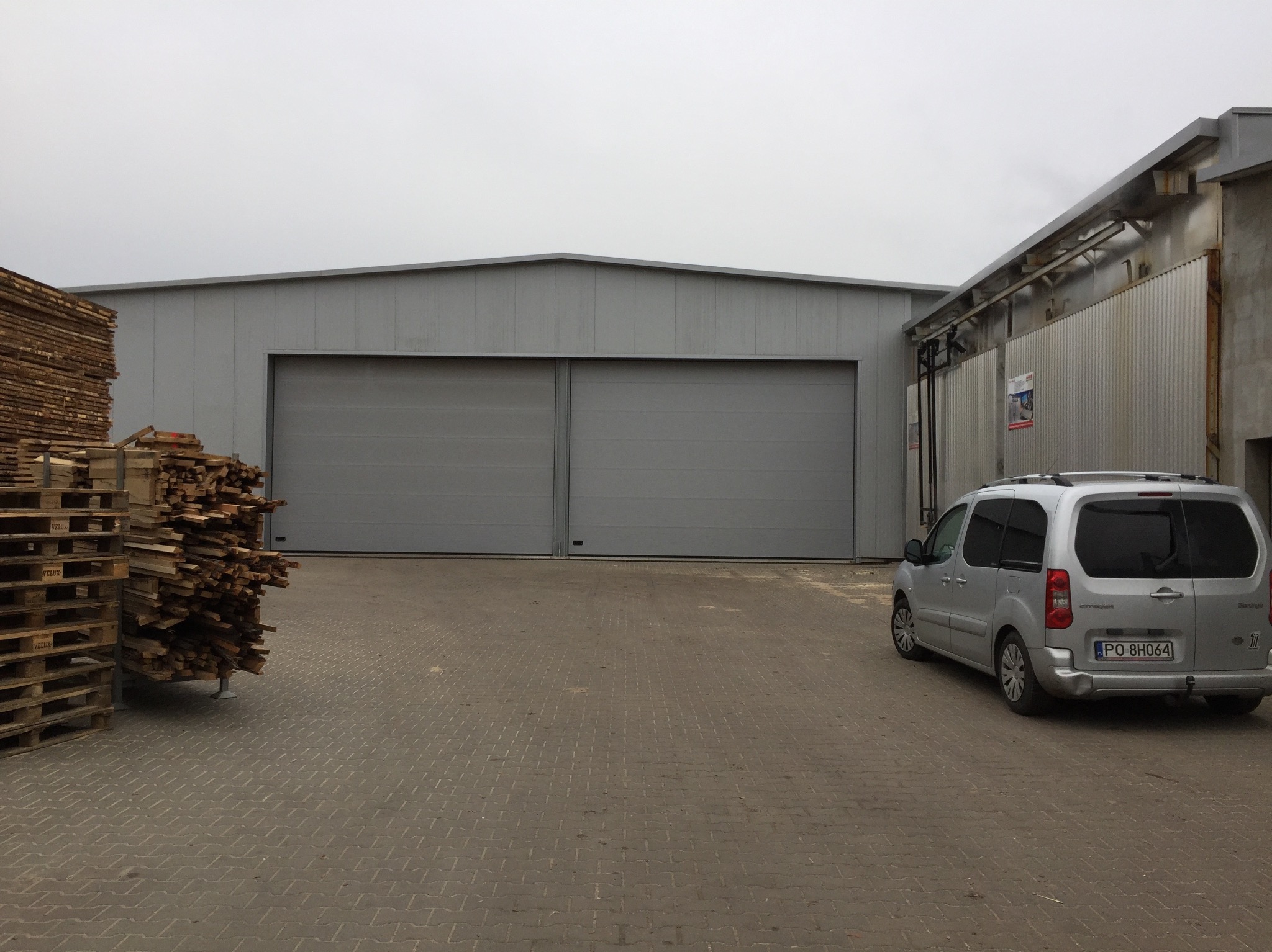 Szara hala magazynowa z dwuskrzydłowymi bramami. Na pierwszym planie palety z drewnem i zaparkowany srebrny samochód dostawczy.