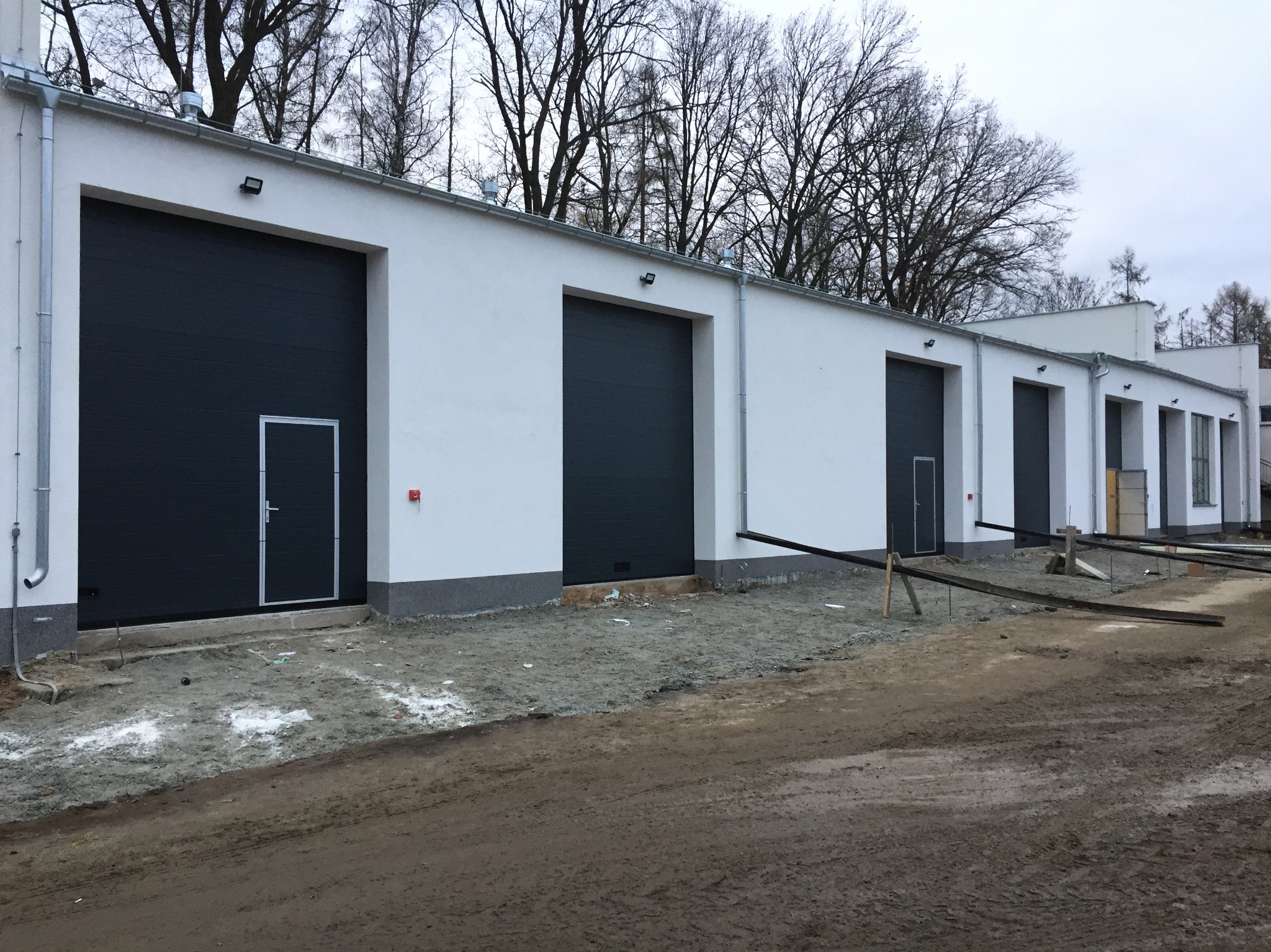 Budynek z szarymi bramami garażowymi segmentowymi i drzwiami wejściowymi. Widoczne elementy orynnowania i instalacji elektrycznej. Tło stanowią drzewa.