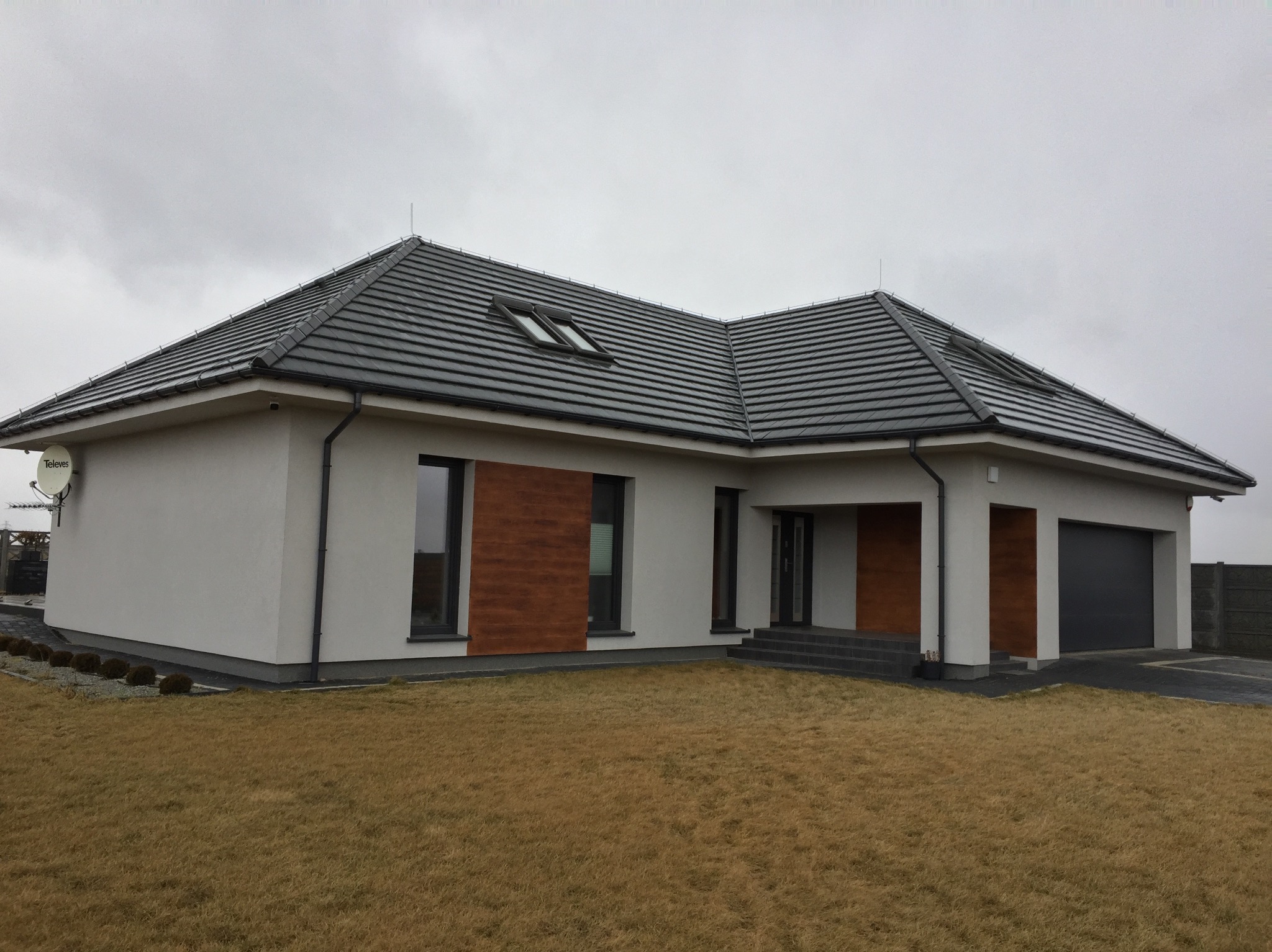 Nowoczesny dom jednorodzinny z grafitową bramą garażową, brązowymi akcentami elewacji i ciemną dachówką pod pochmurnym niebem. Widok z poziomu trawnika.