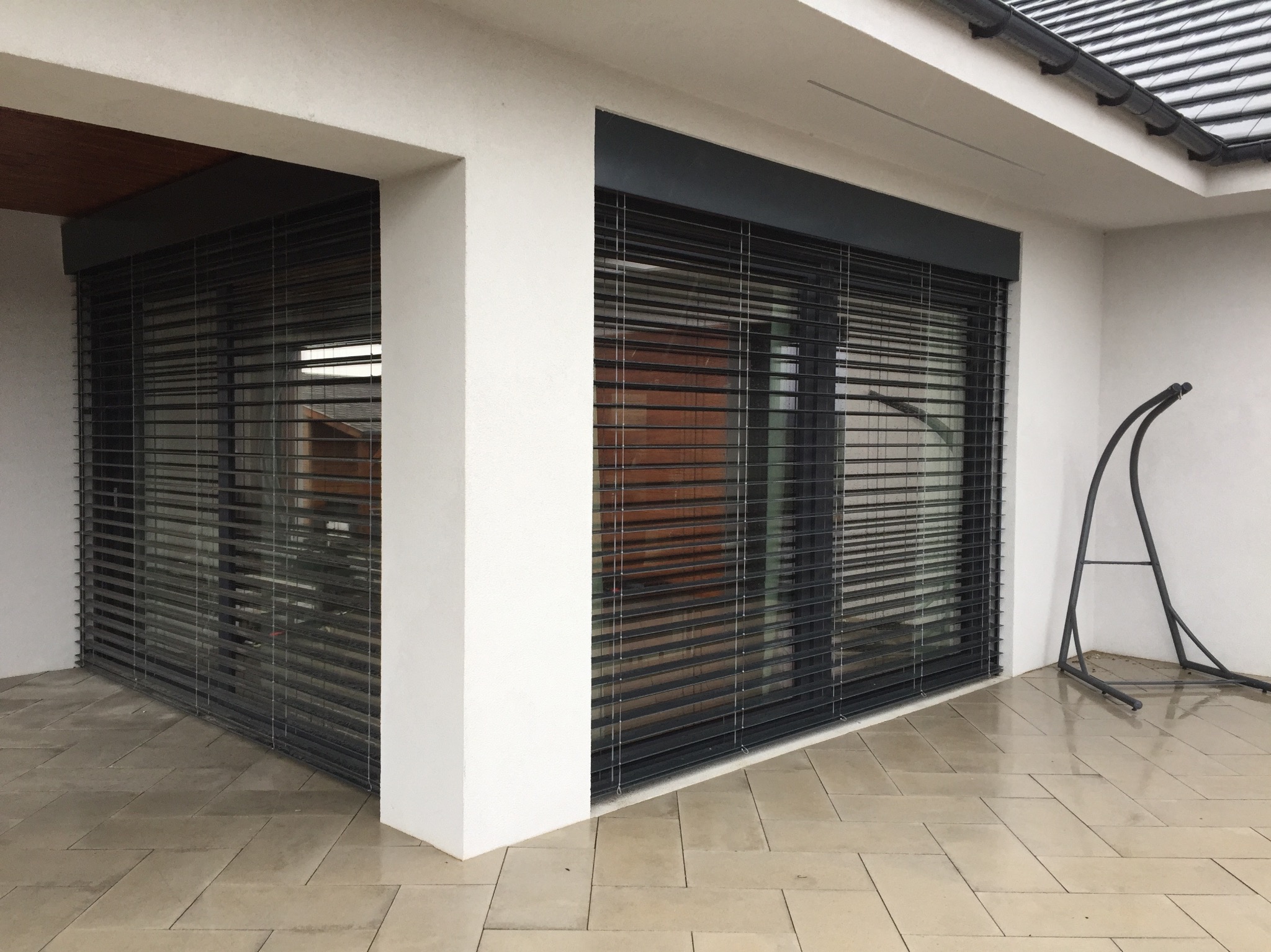 Eleganckie, grafitowe rolety zewnętrzne zamontowane na nowoczesnym budynku z białymi ścianami. Widoczne poziome lamele i fragment tarasu z jasnej kostki.