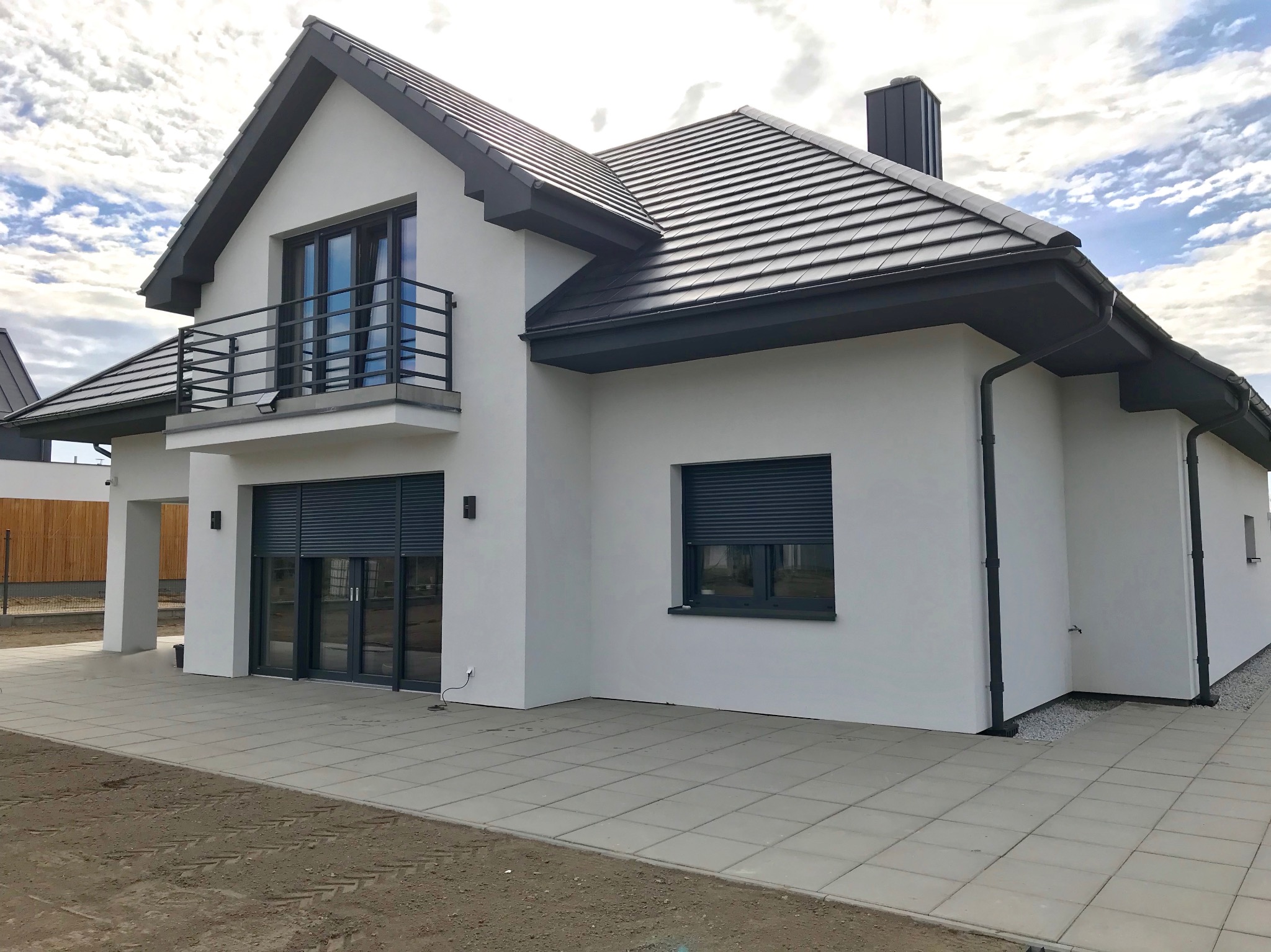 Elewacja domu jednorodzinnego z szarymi roletami zewnętrznymi na parterze i balkonem na piętrze, w tle błękitne niebo z chmurami.