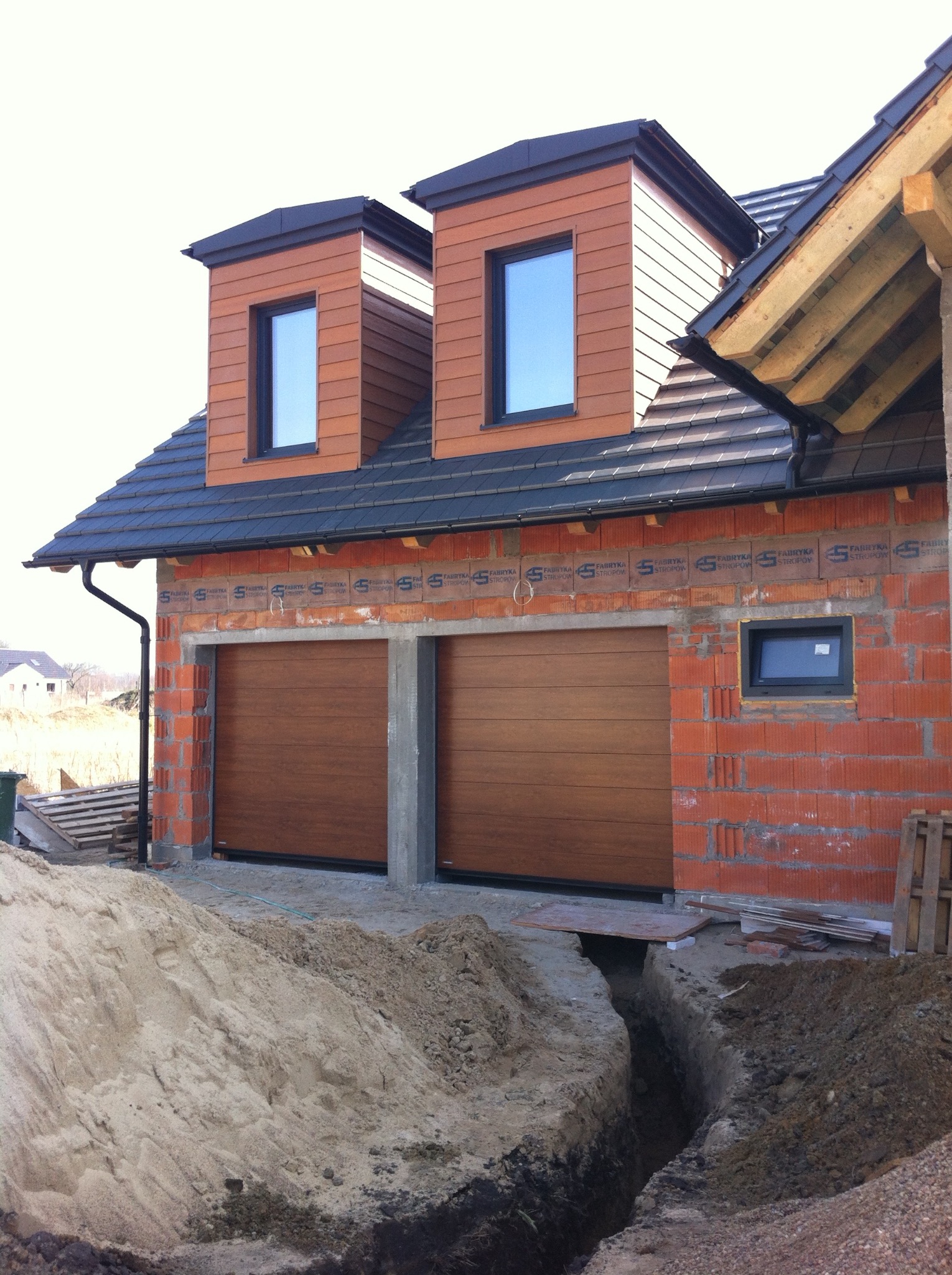 Brązowe, segmentowe bramy garażowe w surowym budynku z cegły, z widocznymi oknami na poddaszu i wykopem przed wjazdem. Dom w trakcie budowy.