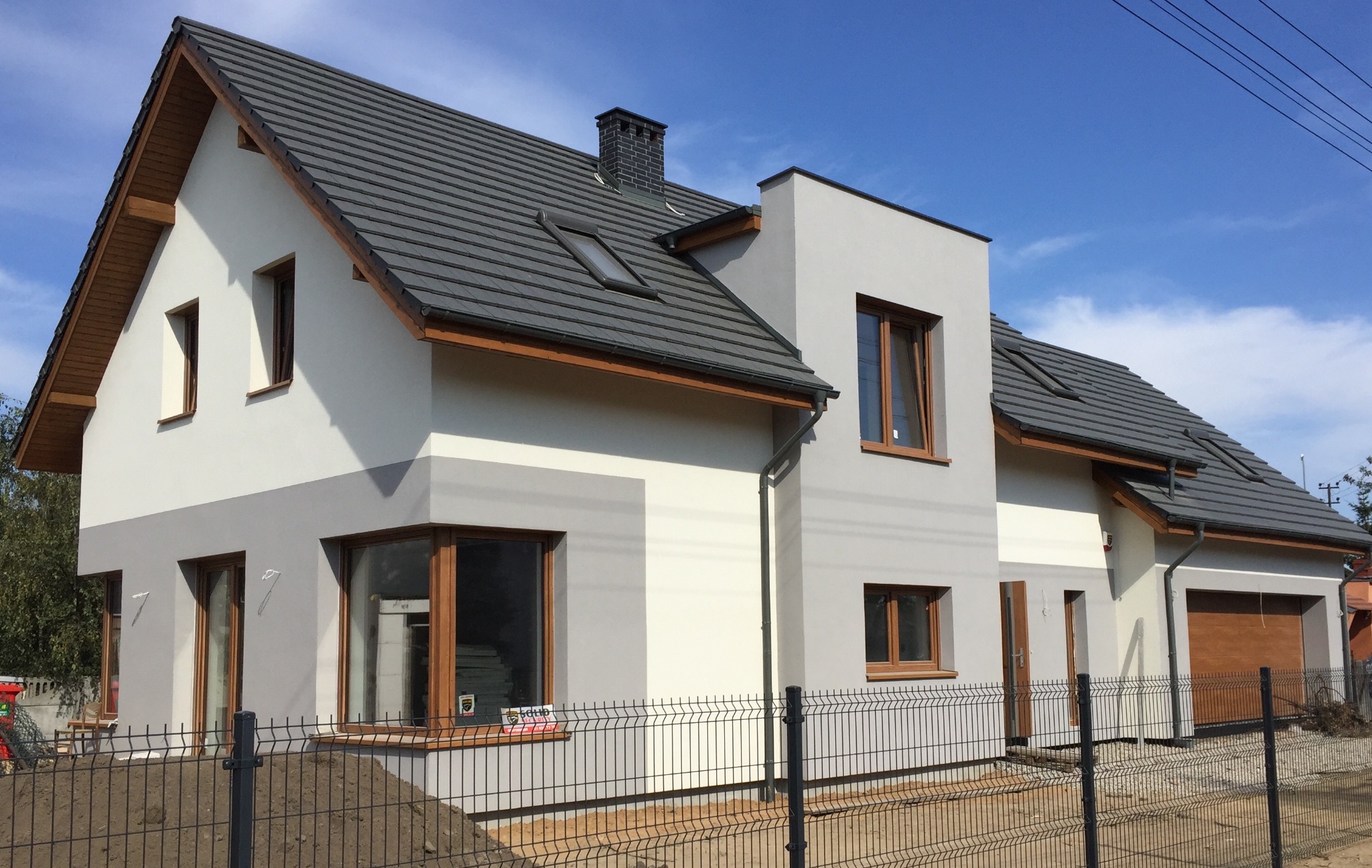 Nowoczesny dom jednorodzinny z grafitową dachówką i drewnianą bramą garażową, otoczony metalowym ogrodzeniem panelowym na tle błękitnego nieba.