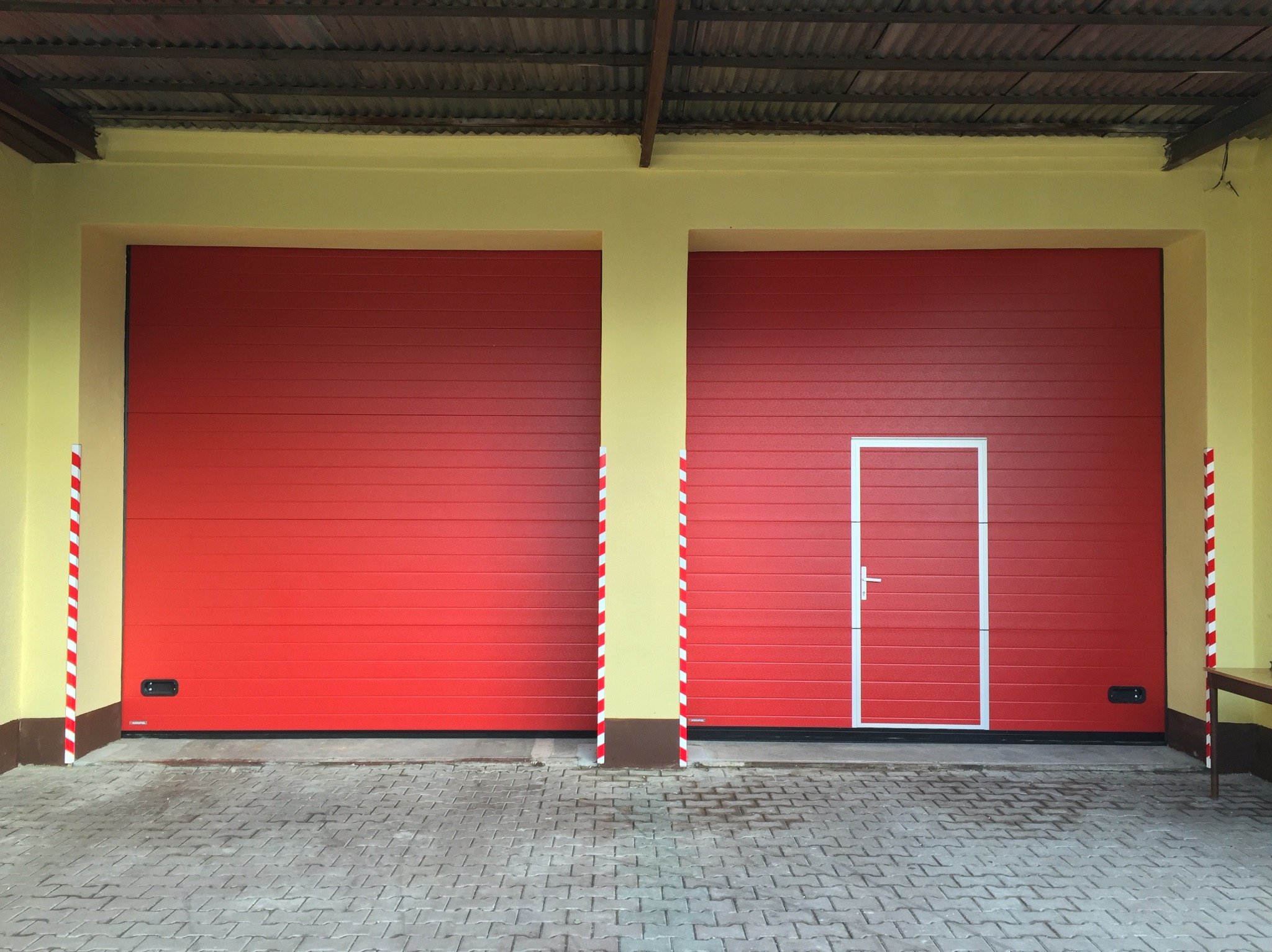 Czerwone bramy garażowe segmentowe z wbudowanymi drzwiami. Kolumny z biało-czerwonymi pasami. Szara kostka brukowa na podjeździe. Żółta elewacja budynku.
