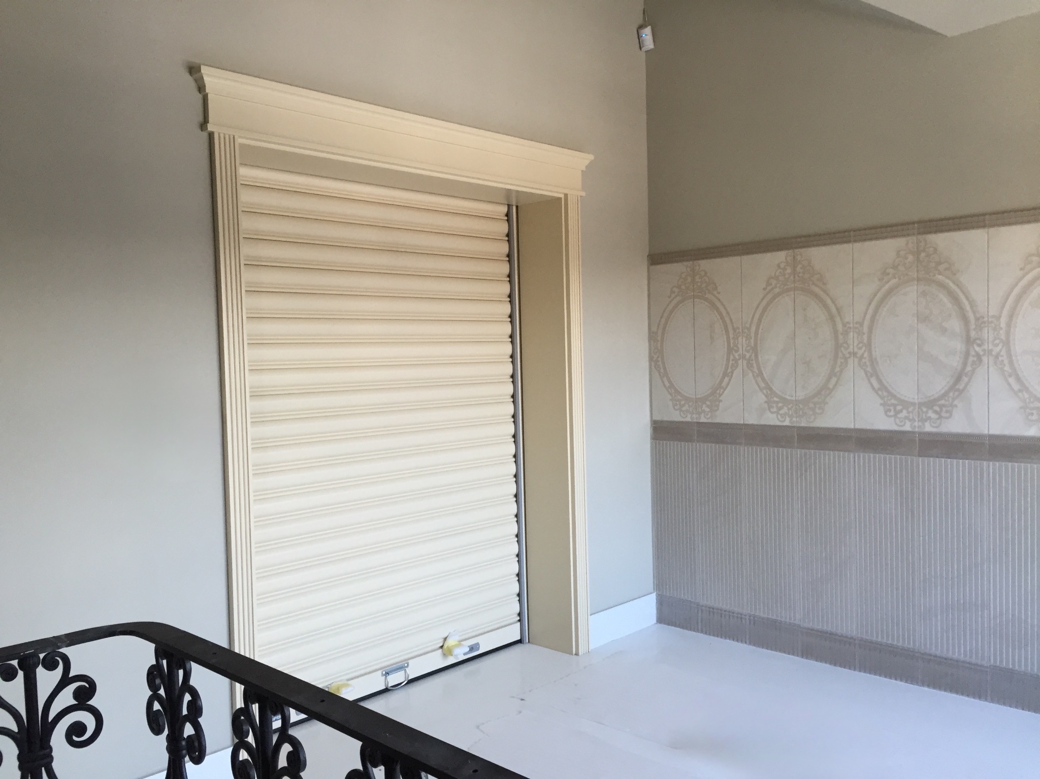 Beżowa brama rolowana w eleganckiej wnęce z dekoracyjną listwą, widoczna z perspektywy klatki schodowej, w jasnym wnętrzu z ozdobnymi płytkami.