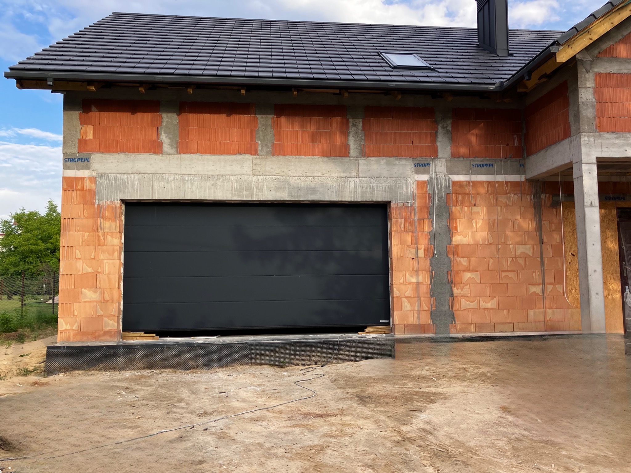 Ciemnoszara brama garażowa segmentowa zamontowana w surowym budynku z czerwonej cegły. Widoczne detale montażowe i fragment dachu z kominem.