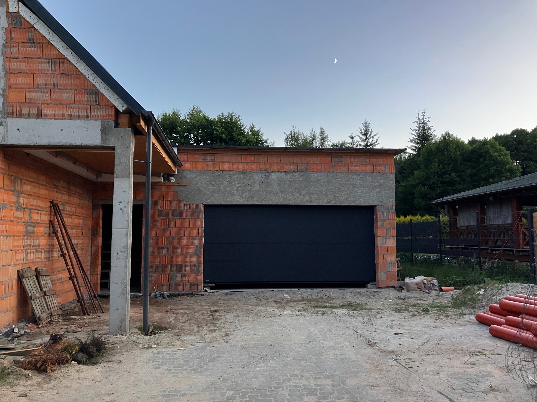 Ciemna brama garażowa segmentowa w surowym otoczeniu budowy z ceglaną elewacją i widocznym księżycem w tle.