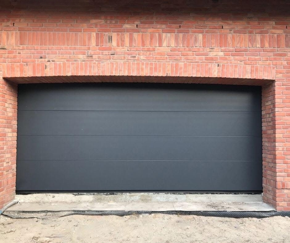 Nowoczesna, segmentowa brama garażowa w kolorze antracytu, osadzona w murze z czerwonej cegły. Brama zamknięta, widoczne poziome przetłoczenia. Surowy beton u dołu.