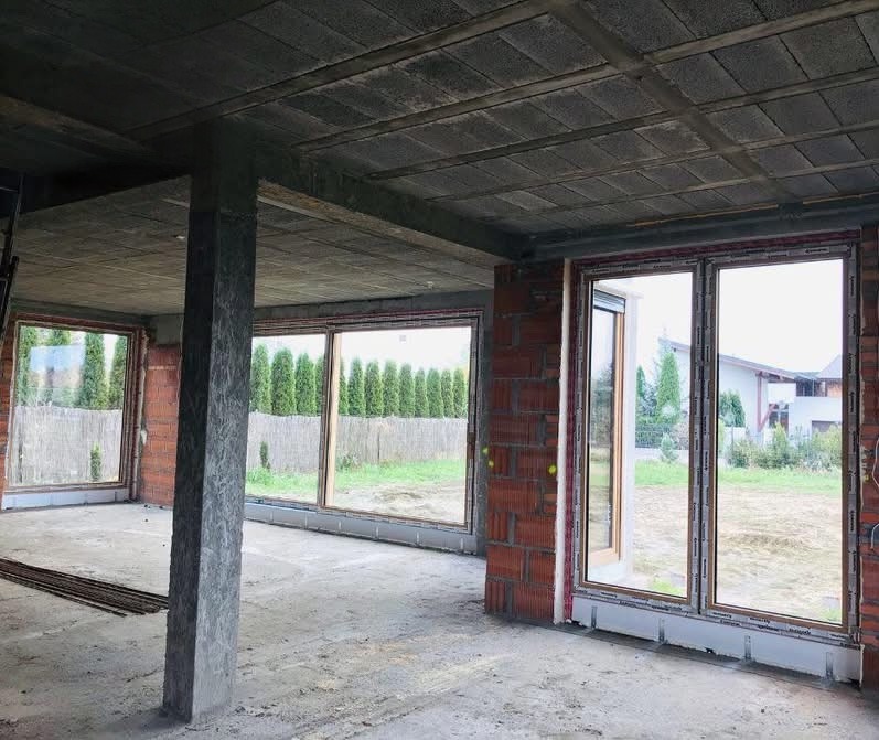 Surowy stan budynku z zamontowanymi oknami PCV, widoczne cegły i betonowy strop. Duże okna z widokiem na zieleń, wnętrze w trakcie budowy.