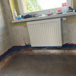 Szymon Kopka - Grzejnik centralnego ogrzewania na tle surowej ściany, widoczne rury instalacji CO w bruzdach podłogi. Na parapecie narzędzia i materiały budowlane.