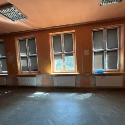 Szymon Kopka - Wnętrze pokoju z trzema oknami i grzejnikami, ściany w odcieniach pomarańczy, podłoga szara. Przy oknie leży niebieska folia. Widoczne ślady po pracach malarskich.