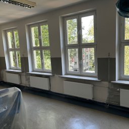 Szymon Kopka - Wnętrze pokoju po malowaniu z trzema oknami i grzejnikami. Świeżo pomalowane ściany w odcieniach szarości i bieli. Podłoga zabezpieczona folią malarską.