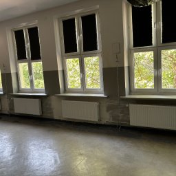 Szymon Kopka - Wnętrze pokoju z trzema oknami z roletami, grzejnikami i szarymi ścianami w trakcie wykańczania. Widoczne rury instalacji grzewczej.