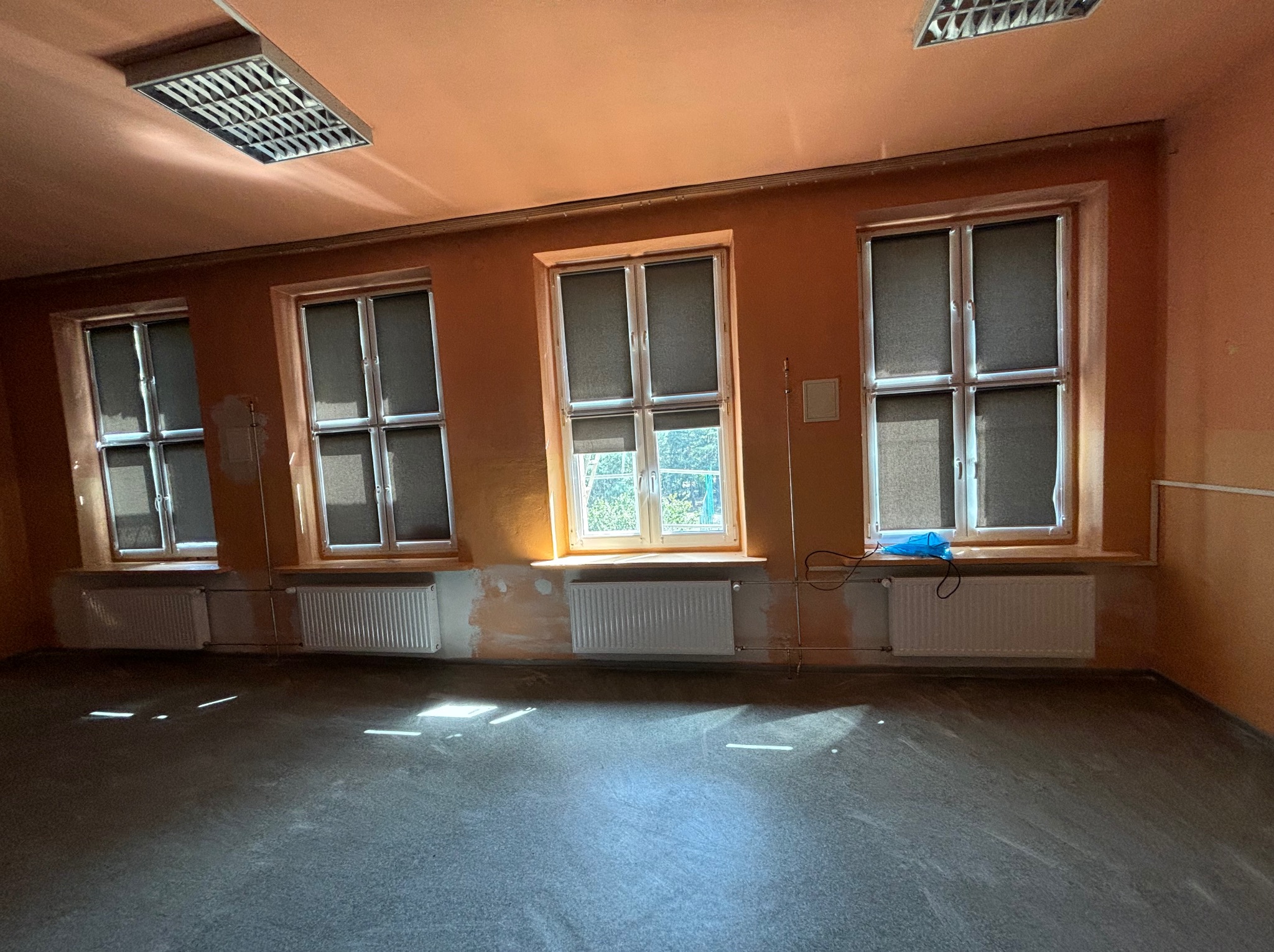 Wnętrze pokoju z trzema oknami i grzejnikami, ściany w odcieniach pomarańczy, podłoga szara. Przy oknie leży niebieska folia. Widoczne ślady po pracach malarskich.