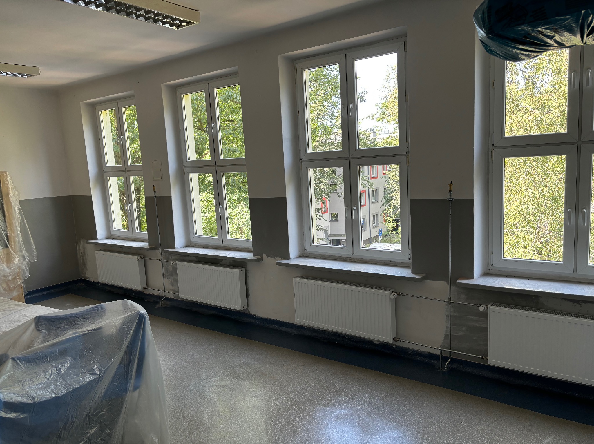 Wnętrze pokoju po malowaniu z trzema oknami i grzejnikami. Świeżo pomalowane ściany w odcieniach szarości i bieli. Podłoga zabezpieczona folią malarską.