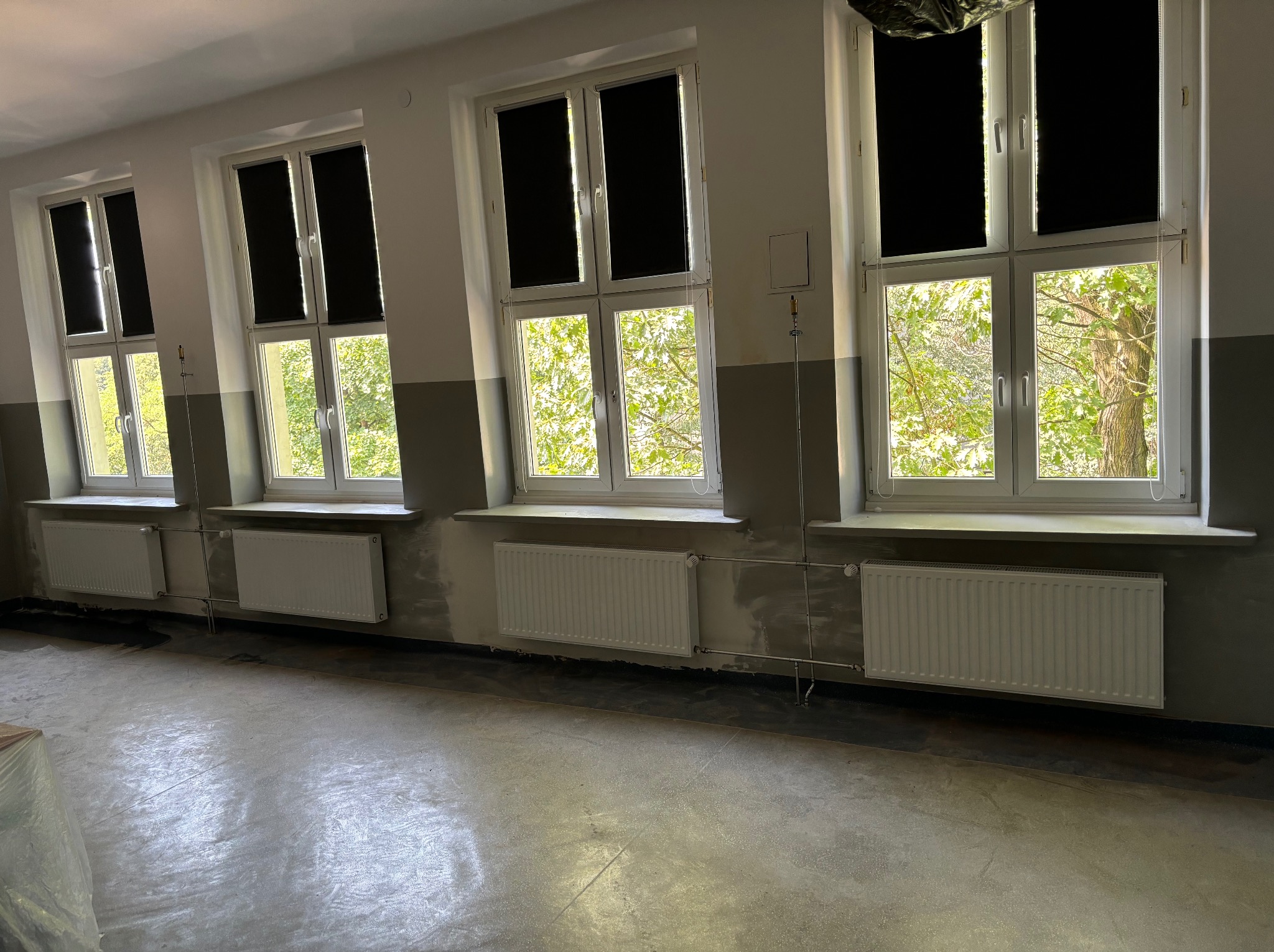 Wnętrze pokoju z trzema oknami z roletami, grzejnikami i szarymi ścianami w trakcie wykańczania. Widoczne rury instalacji grzewczej.