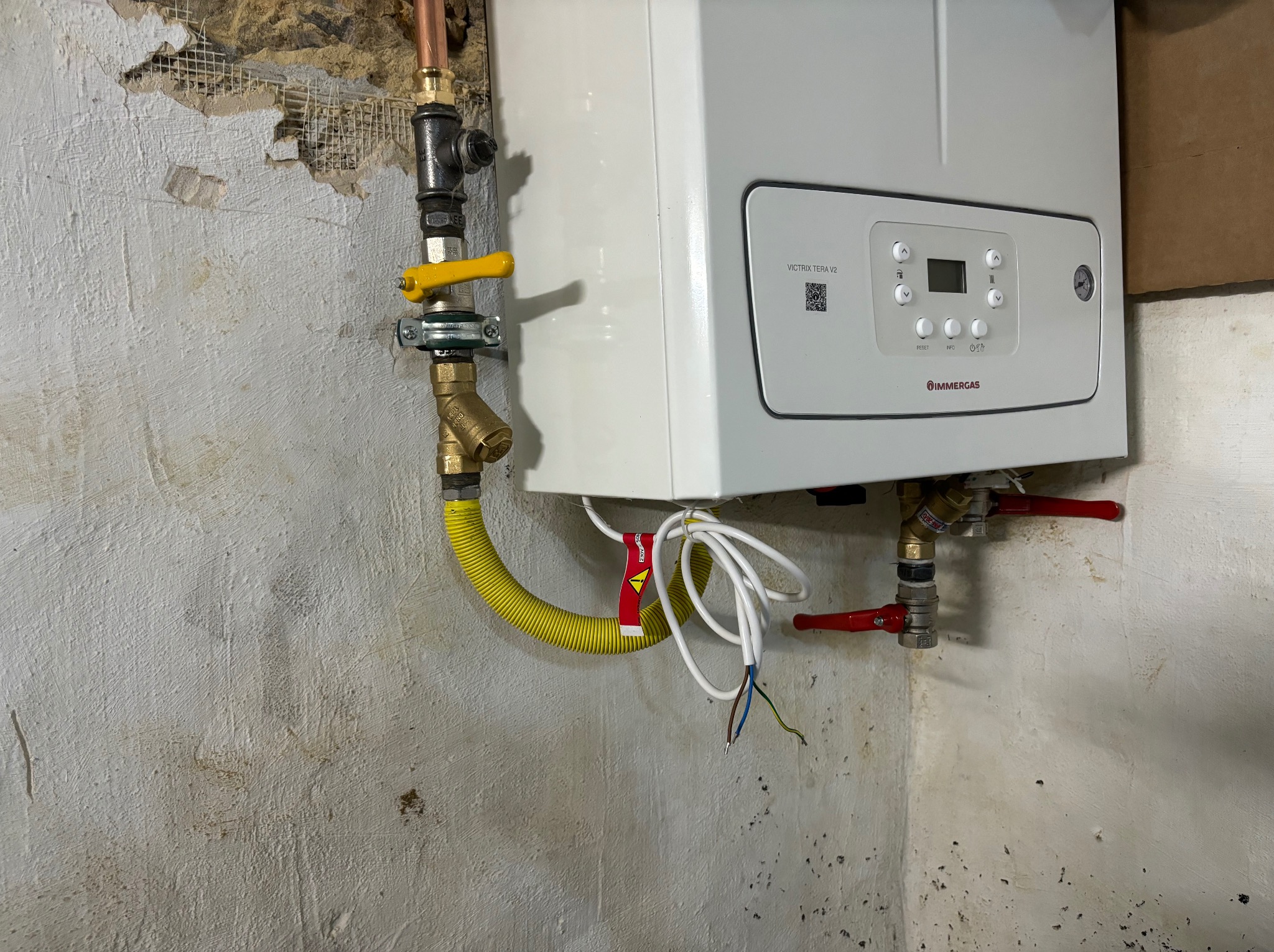 Biały piec gazowy Immergas Victrix Tera V2 zamontowany na ścianie, z widocznymi przyłączami gazowymi i elektrycznymi. Ściana w tle z widocznymi śladami remontu.