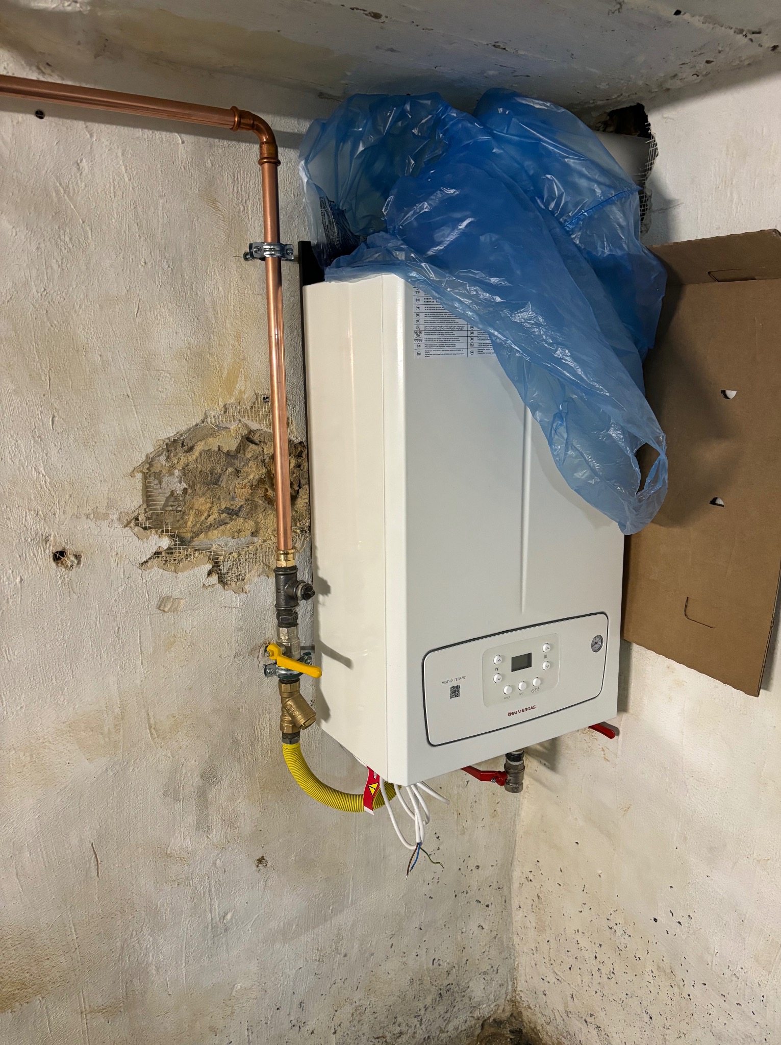 Biały piec gazowy Immergas, zamontowany na ścianie z widocznymi śladami napraw, z podłączonymi miedzianymi rurami i żółtym przewodem gazowym. Częściowo osłonięty niebieską folią.