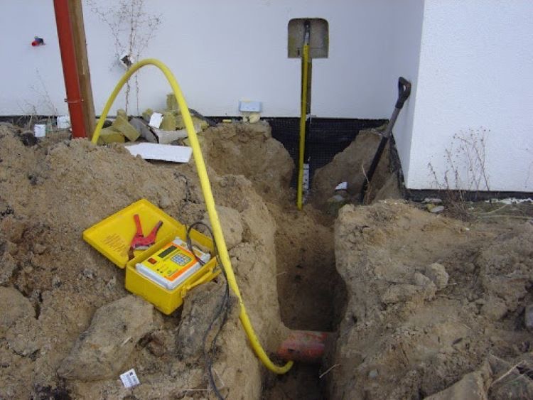 Wykop z żółtą rurą gazową, miernikiem w żółtej obudowie i łopatą. Widoczna ściana budynku z izolacją. Prace ziemne podczas instalacji gazowej.