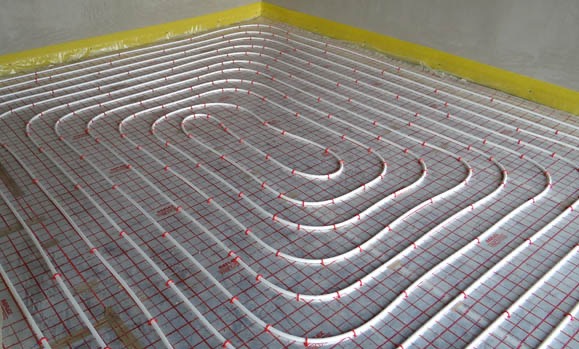 Ułożone rury ogrzewania podłogowego na siatce z czerwonymi znacznikami, zabezpieczone żółtą folią przy ścianie, gotowe do zalania wylewką.