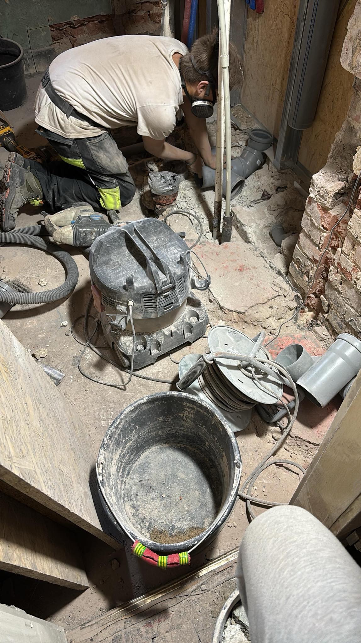 Instalator w masce łączy rury kanalizacyjne w piwnicy. W tle widoczne narzędzia, wiadro i odkurzacz przemysłowy. Surowy stan pomieszczenia przed wykończeniem.