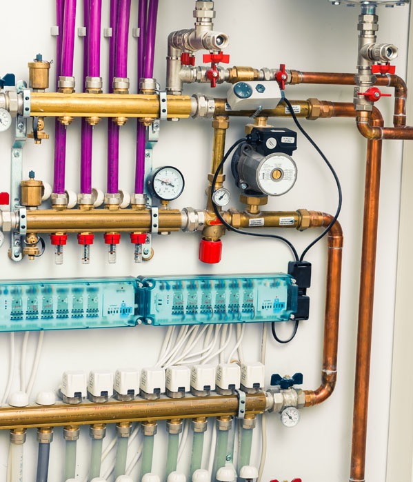 Rozdzielacz ogrzewania podłogowego z purpurowymi rurami, manometrami i zaworami na białej ścianie. Widoczne miedziane rury i elementy sterujące.