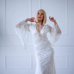 Tamka Foto Studio - Blond modelka w koronkowej sukni z rozszerzanymi rękawami prezentuje stylizację na tle białej ściany z panelami. Całość dopełnia perłowa biżuteria.